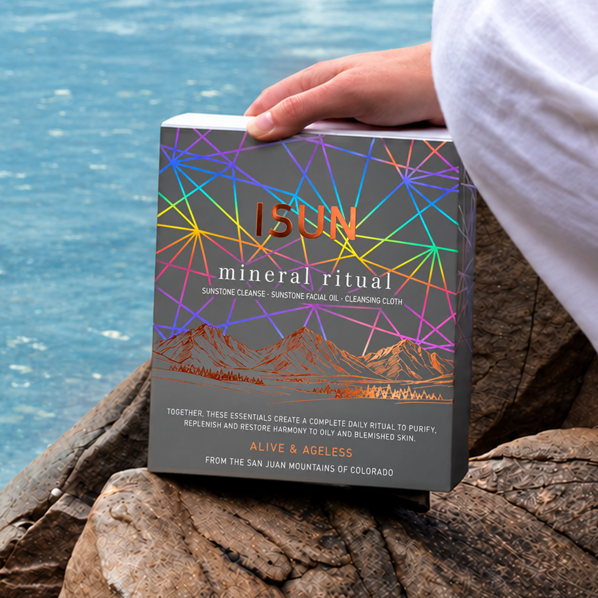 En person holder en ISUN Mineral Ritual-gaveeske med geometriske linjer på steiner ved vannet.