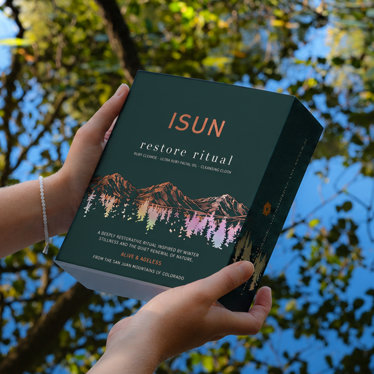 Hender som holder en ISUN Restore Ritual-gaveeske ved vann som speiler grønt løvverk.