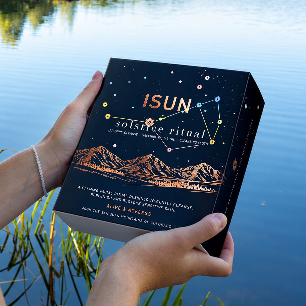 Hender som holder en ISUN Solstice Ritual-gaveeske med stjerner og fjellmotiv ved en innsjø.