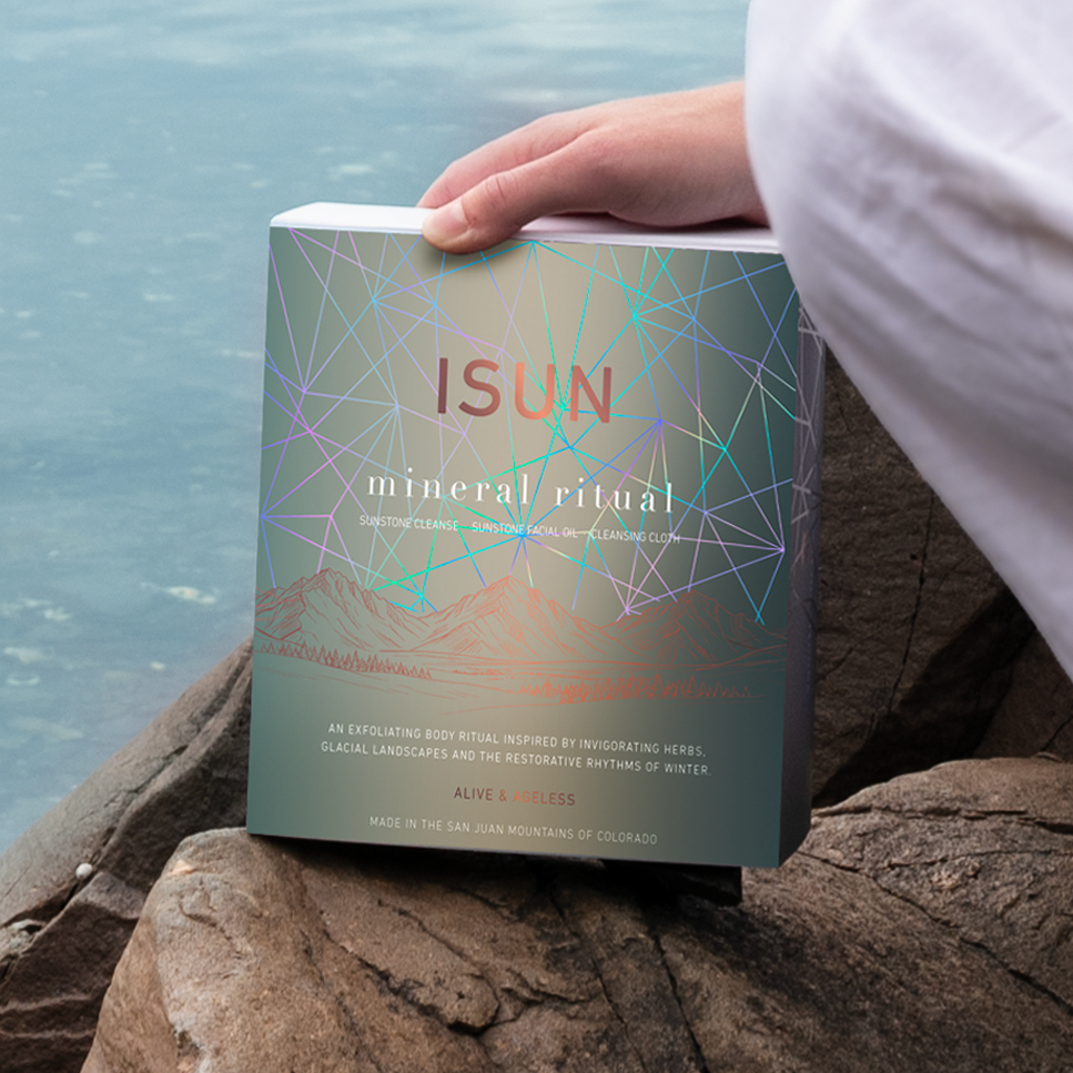 En person holder en ISUN Mineral Ritual-gaveeske med geometriske linjer på steiner ved vannet.