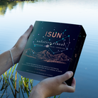 Hender som holder en ISUN Solstice Ritual-gaveeske med stjerner og fjellmotiv ved en innsjø.